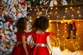 Two curly little girls looking at the christmas fireplace near beautiful Christmas tree. Twins in red dresses looking at Royalty Free Stock Photo