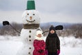 Two cute children, boy and girl, standing in front of smiling snowman in bucket hat, scarf and gloves on snowy winter landscape an Royalty Free Stock Photo