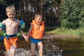 two cute little boys running into lake splashing water in forest. one boy wearing life vest, another wearing armbands Royalty Free Stock Photo