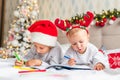 Two cute little girls sisters wearing Santa Helper hats write letter to Santa lying on bed at home in Christmas