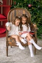 Two cute little sisters are sitting in a chair by a Christmas tree decorated with lights and reading xmas fairy tales.