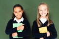two cute multicultural schoolgirls smiling at camera while standing near chalkboard and holding books.