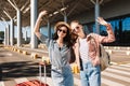 Two cute smiling girls in sunglasses happily looking in camera while raising hands up with suitcase and backpack on