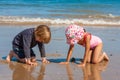 Two Cute Young Children kneeling playing in the sand together on a beach by the seaside