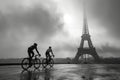 Two cyclists ride before Eiffel Tower's silhouette in fog.