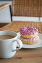 Donuts with pink icing and a cup of coffee with milk on a wooden table against the background of a chair Royalty Free Stock Photo