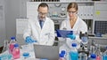 Two eager scientists engrossed in document analysis on a laptop at their bustling lab, amongst test tubes, microscopes and