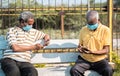 Two elderly people with medical face mask busy using mobile phone while sitting at park while maintaining social distance -