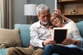 Two elderly people senior couple man and woman looking at a family photo smile and hug while sitting at home on sofa. memories and Royalty Free Stock Photo