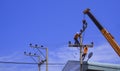 Two electricians with crane truck are installing electrical equipment on power pole against blue sky