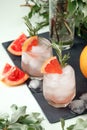 Two elegance cold wet drinking glasses with fresh grapefruit summer cocktail, ice, rosemary on back stone background and greenery