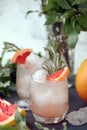 Two elegance cold wet drinking glasses with fresh grapefruit summer cocktail, ice, rosemary on back stone background and greenery