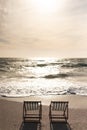 Two empty folding wooden chairs on shore in front of waves splashing in sea at beach against sky