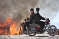 Two boys enjoying on motorcycle while woods stalls are burning.