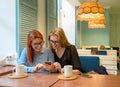 Two excited young girls using mobile phones, sitting in a cafe and pointing finger. Red-haired woman in glasses shows Royalty Free Stock Photo