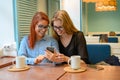Two excited young girls using mobile phones, sitting in a cafe and pointing finger. Red-haired woman in glasses shows Royalty Free Stock Photo