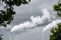 Two factory smokestacks emitting white smoke against a stormy sky with foliage in the foreground Royalty Free Stock Photo