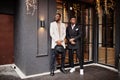 Two fashion black men pose against house with garlands. Fashionable portrait of african american male models. Wear suit, coat and Royalty Free Stock Photo