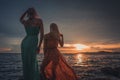 Two fashion models posing in red and turquoise dresses in the rays of the setting sun on a tropical beach. Back to the Royalty Free Stock Photo