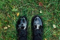 Two feet in black shoes stepping on the green grass with fallen autumn leaves.