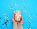 Two female hands holding a brown box with a pink bow on a blue background with multi-colored confetti Royalty Free Stock Photo