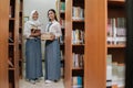 two female students in high school uniforms carry several books standing between the bookshelves Royalty Free Stock Photo