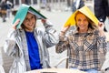 Two female students sitting outside at a terrace table covering their heads for the rain with two colored plastic