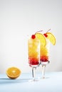 Two festive cocktails of tequila Sunrise on a blue and white background, next to a slice of orange.