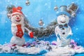 Two festive snowmen on blue background. It`s snowing around, Christmas balls hang and lie