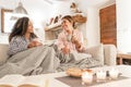 Two fine young women sitting on the sofa with a blanket on their legs laughing enjoying the house in winter having tea with