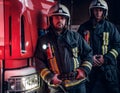 Two firemen wearing protective uniform standing next to a fire truck in a garage of a fire department. Royalty Free Stock Photo