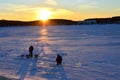 Two fishermen on a frozen lake catch fish in a hole of ice on the background of evening sunset. Winter arrives, bodies of water