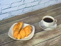 Two freshly baked croissants on a white ceramic plate and a cup of black coffee on a saucer with a spoon on a wooden table against Royalty Free Stock Photo