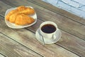 Two freshly baked croissants on a white ceramic plate and a cup of black coffee on a saucer with a spoon on a wooden table against Royalty Free Stock Photo