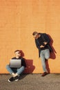 Two friends in casual-wear in urban contest with digital devices. Young caucasian man sitting cross-legged on ground and working Royalty Free Stock Photo