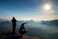 Two friends. Hiker thinking and photo enthusiast takes photos stay on cliff. Dreamy fogy landscape, blue misty sunrise in a beaut