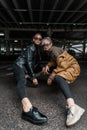 Two friends pretty young girls in fashion cool clothes with a leather jacket and black jeans posing in a parking lot in the city