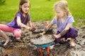 Two funny little girls playing in a large wet mud puddle on sunny summer day. Children getting dirty while digging in muddy soil