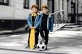 Two funny twin boys posing with happy faces in the street. Skateboard or pennyboard and soccer ball.