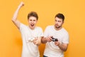 Two gamers in white T-shirts hold the joystick in their hands and emotionally play video games, isolated on a yellow background.