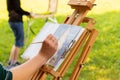 Artist painting on open air, wooden easel sketchbook with unfinished painting close-up. Female hand paints in a sunny summer Royalty Free Stock Photo