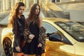 Two girls on a background of a yellow car. Beautiful brunettes near the cottage arrived on an expensive car. Famous girls.