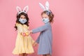 Two girls in bunny ears headband and protective mask with colored eggs on pink background. Covid easter concept