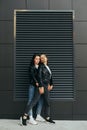 Two girls in dark casual clothes stand on a black background, hugging and posing at the camera.Attractive best girlfriends are