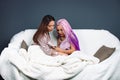 Two girls at home wrapped in a white fluffy blanket, watching social news in pandemic on a smartphone. Gray background
