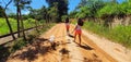 Two girls with long hair, pink shorts and a white shirt, walk with a white poodle named Nina, along the dirt road, flanked by