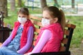 Two girls in medical masks sit on a bench in a park