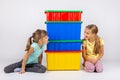 Two girls peek from different sides of large plastic boxes and show each other their tongues