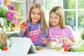 Two girls in pink aprons preparing salad on kitchen table with tablet