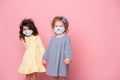 Two girls in protective mask with colored eggs holding hands on pink background. Covid easter concept Royalty Free Stock Photo